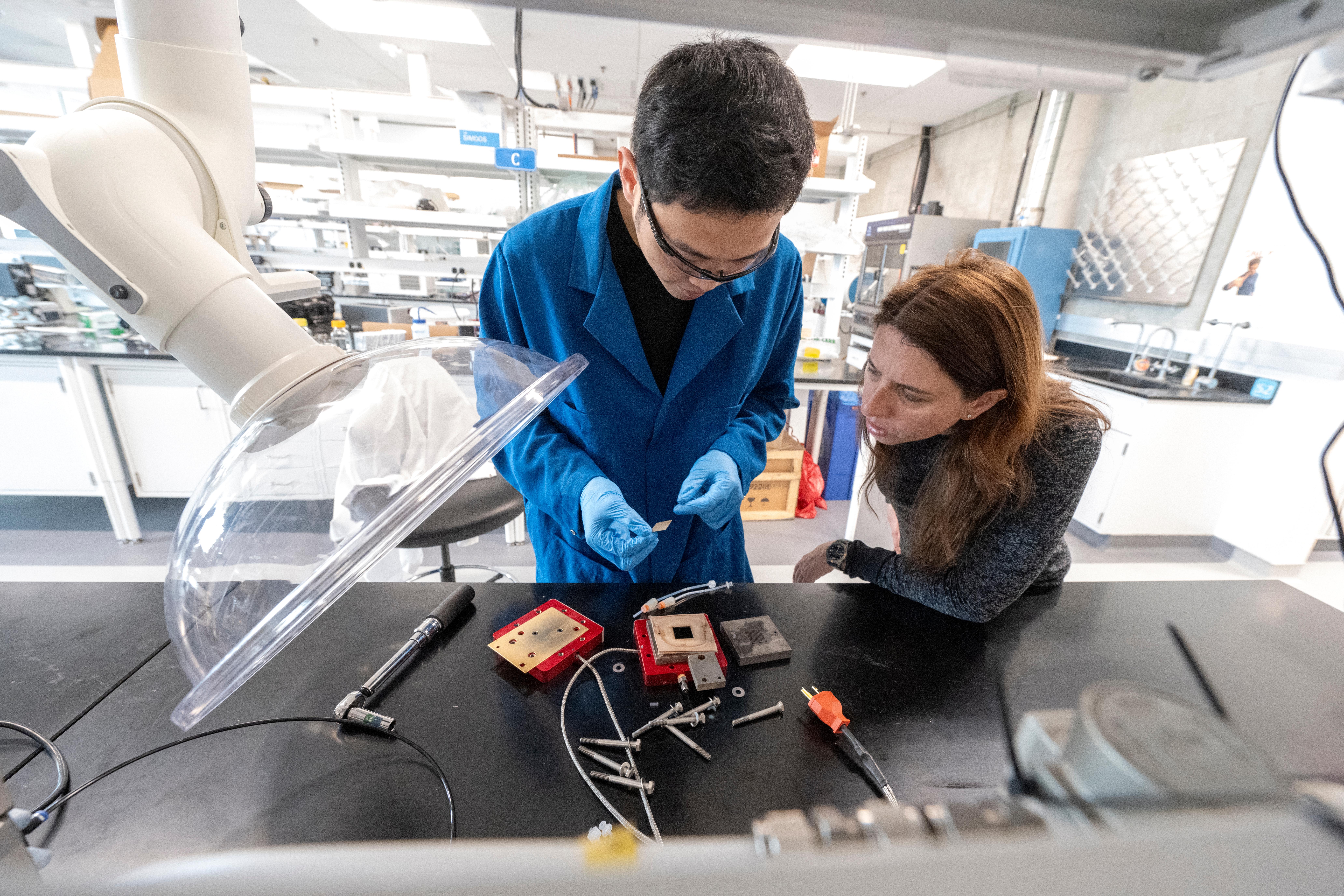 A student wearing a blue lab coat and gloves examines a small sample while a researcher observes beside them in a laboratory. Scientific instruments and tools are arranged on the workbench.
