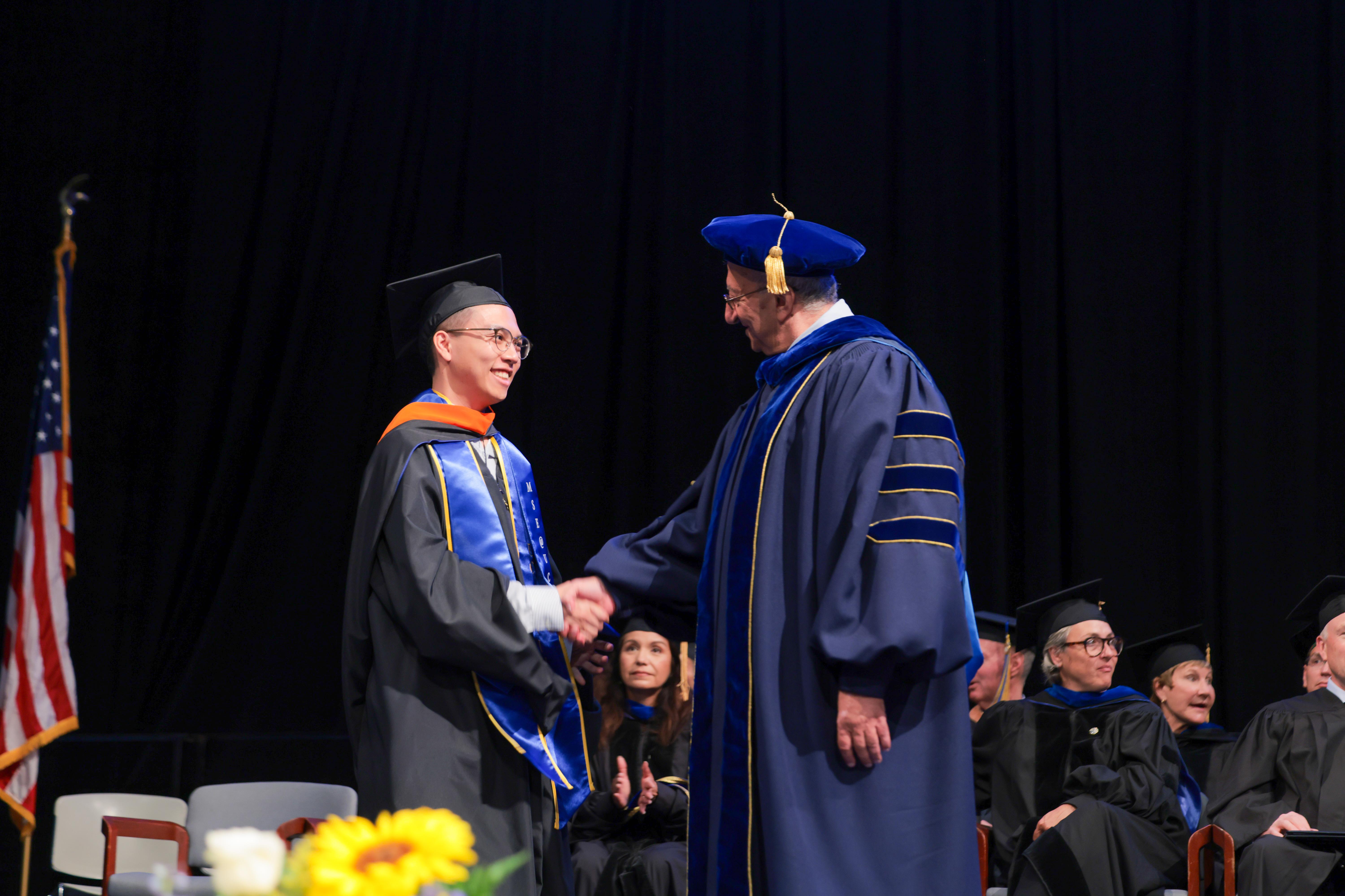 A graduate in academic regalia is shaking hands with a professor, also in academic regalia, on a stage during a graduation ceremony. Other graduates and faculty members are visible in the background.