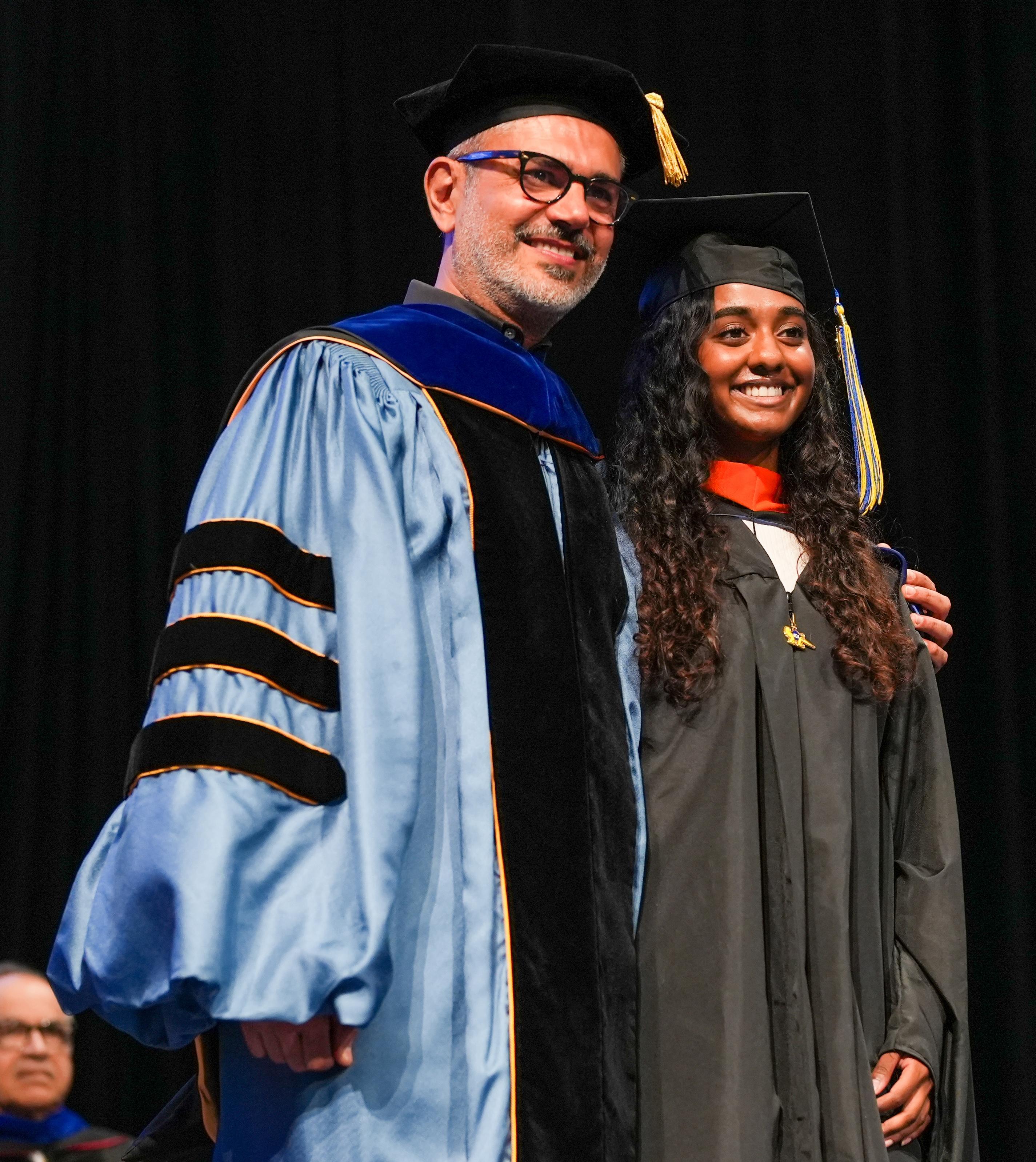 A faculty member wearing blue and black academic regalia stands with a smiling graduate in a black cap and gown on stage during a commencement ceremony.