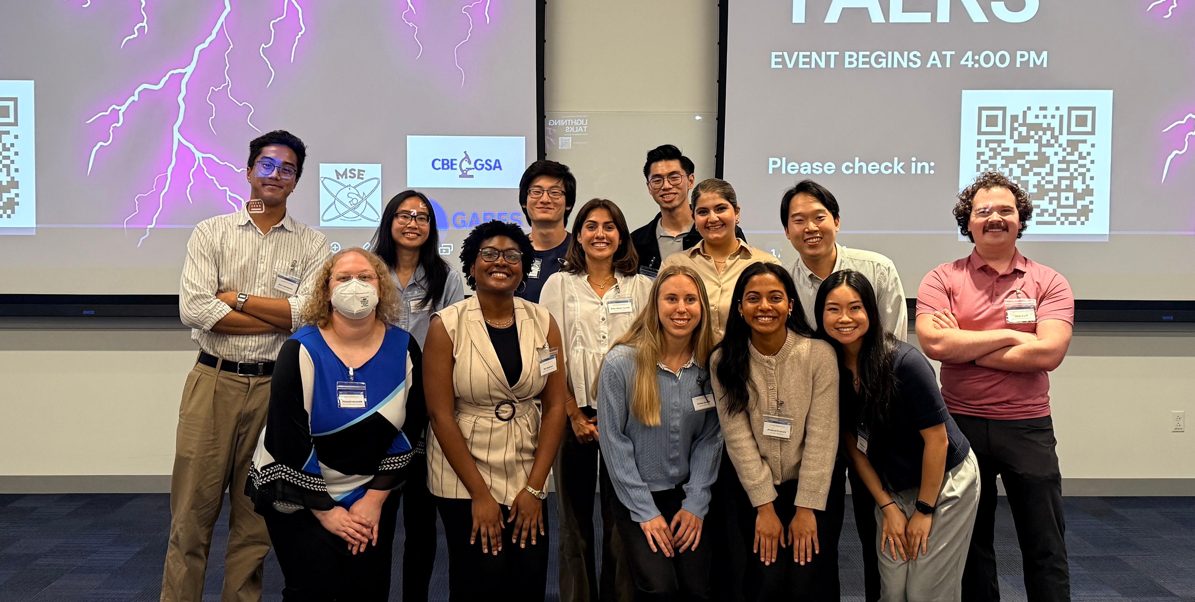 Thirteen students and organizers stand together in a lecture hall, smiling for a group photo in front of two large presentation screens displaying the "Lightning Talks" event title and a QR code for check-in.