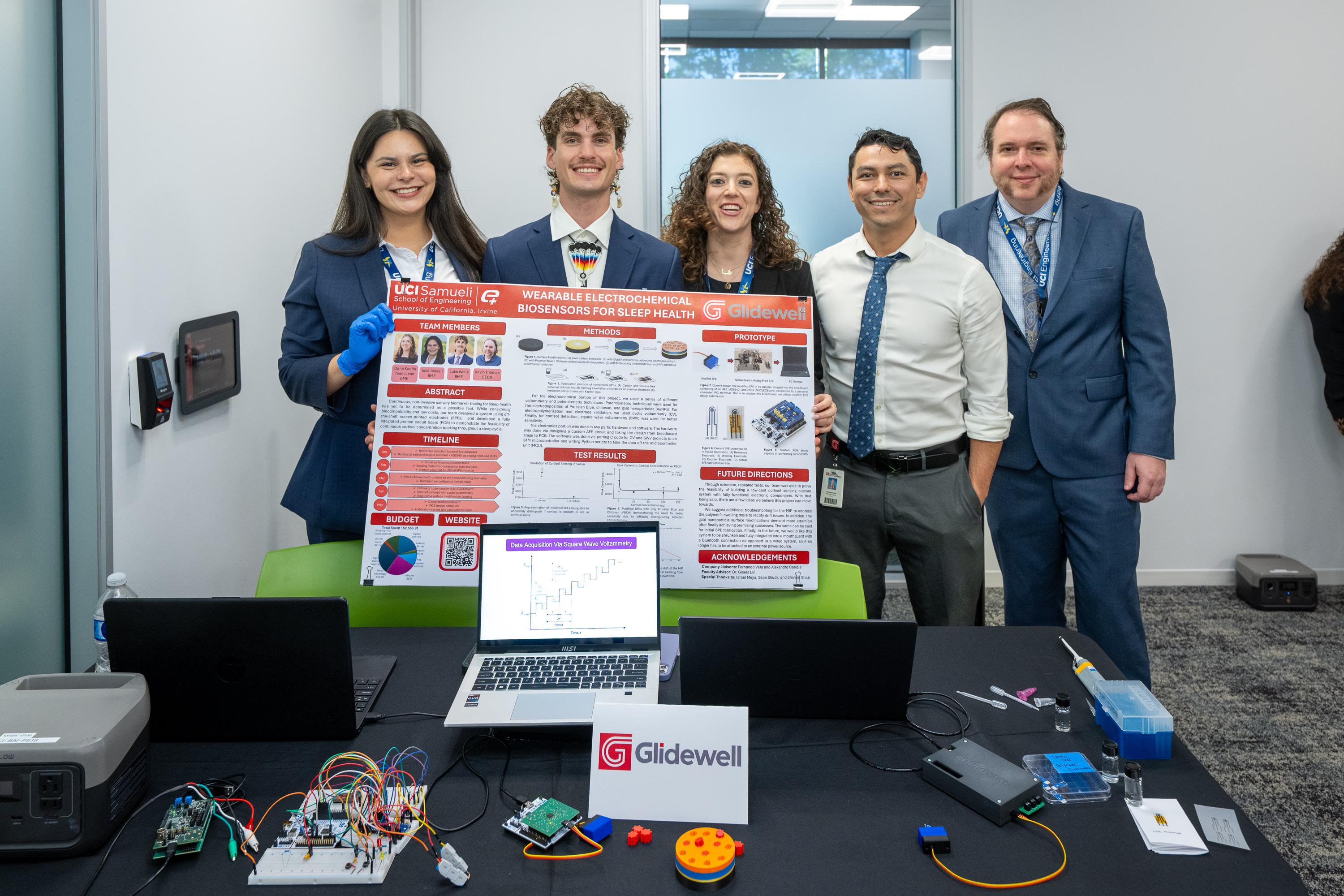 Master of Engineering capstone team presenting their project, "Wearable Electrochemical Biosensors for Sleep Health," alongside faculty and an industry mentor at the UC Irvine Capstone Showcase. A project poster, laptop, and prototype components are displayed on the table.