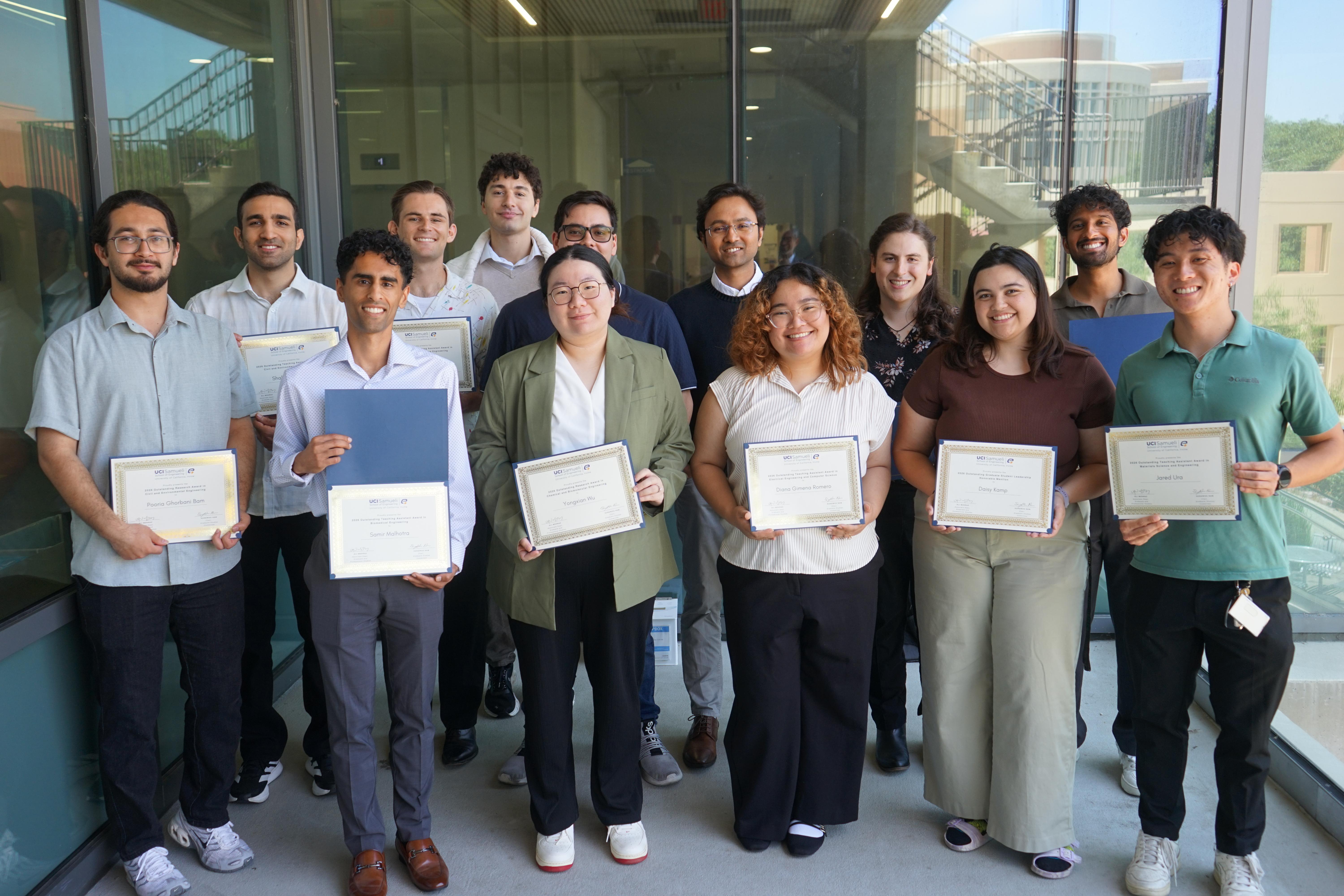 Fourteen students pose together holding certificates of recognition in a campus building. The group smiles toward the camera, with several students standing in front and others behind them.