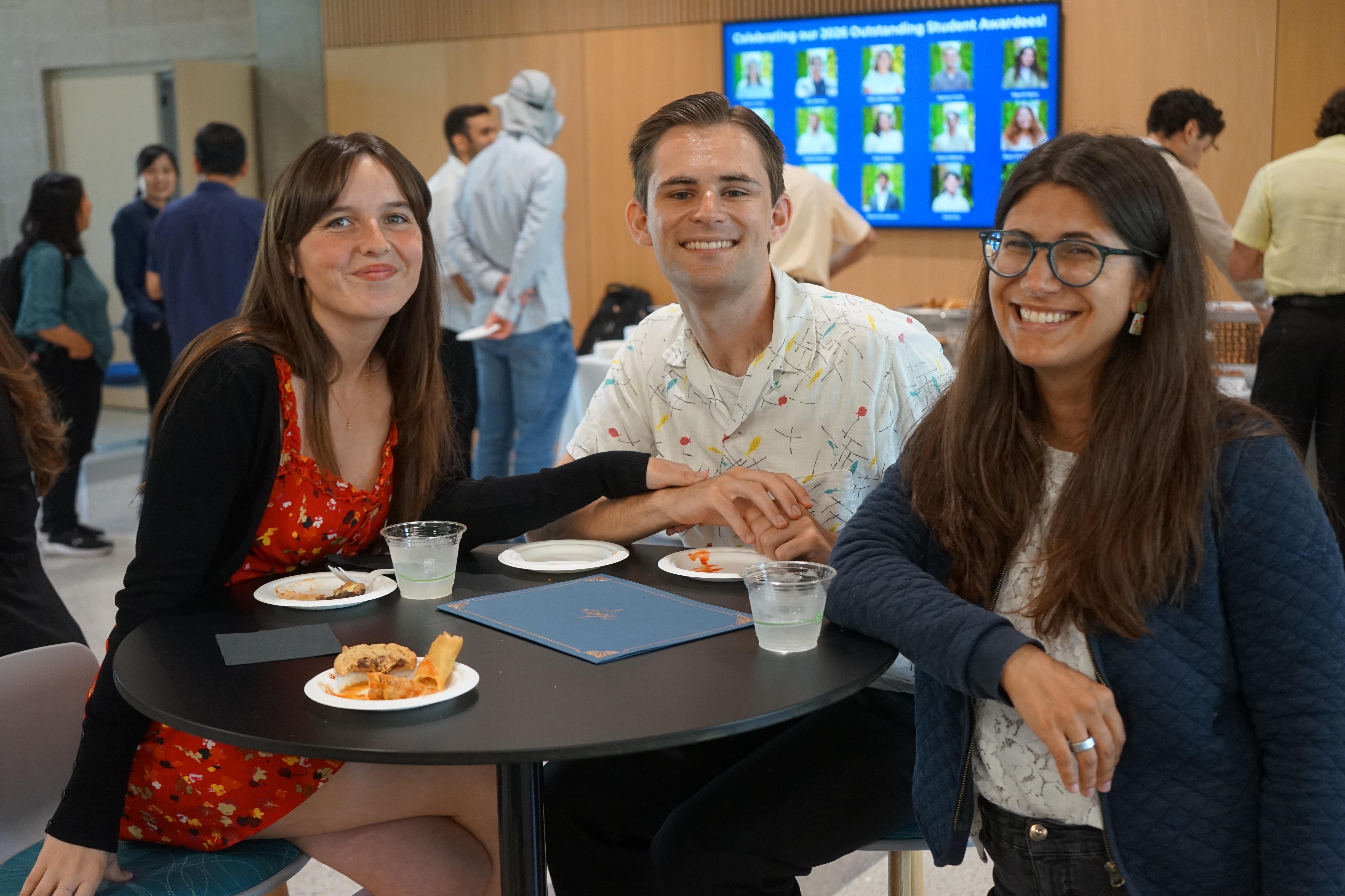 Three smiling individuals are seated around a table, with food and drinks present. In the background, a large screen displays images and text related to student awards.