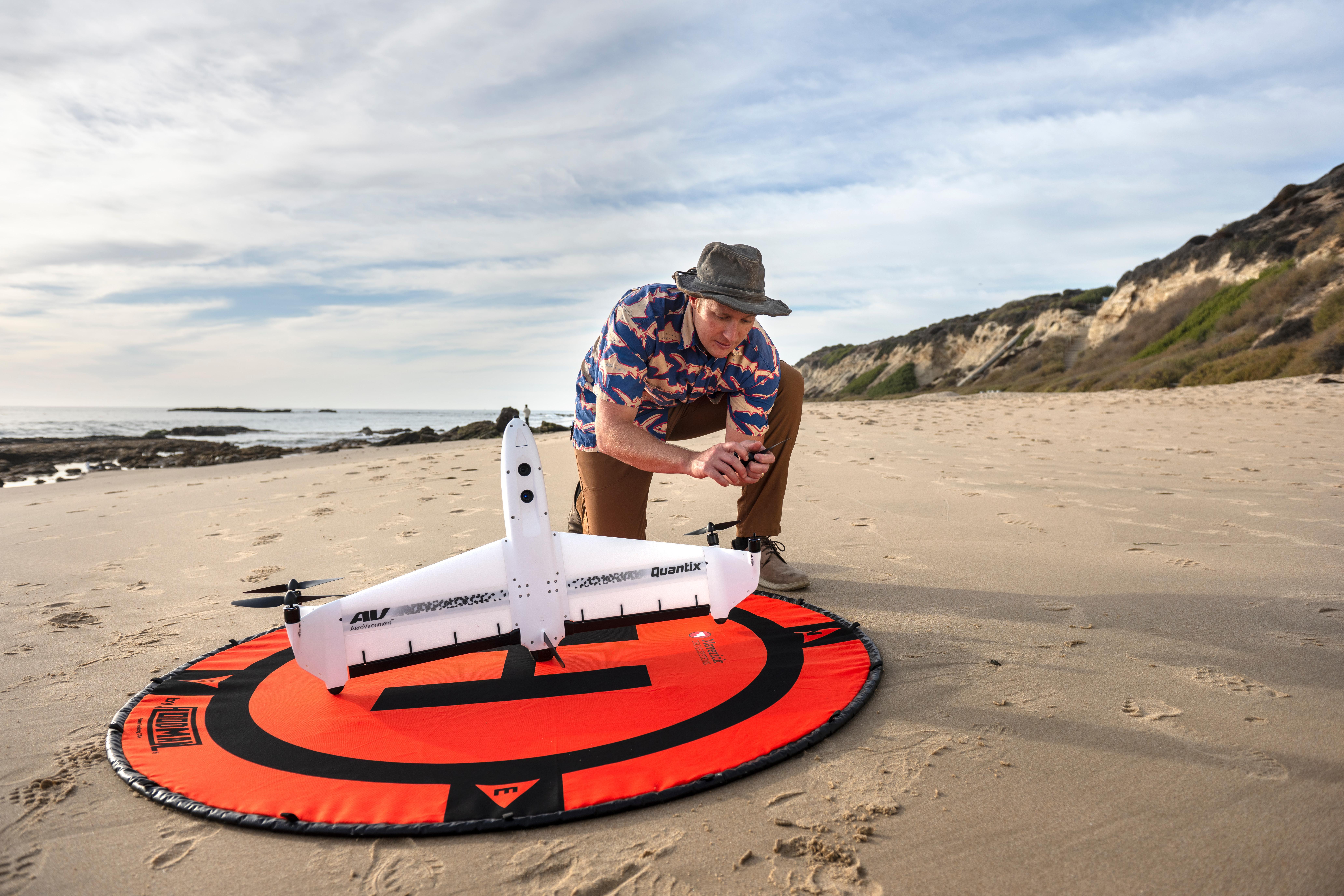 A researcher kneels on a beach while preparing a fixed-wing drone on a landing pad before a coastal field survey. Ocean waves and cliffs are visible in the background.